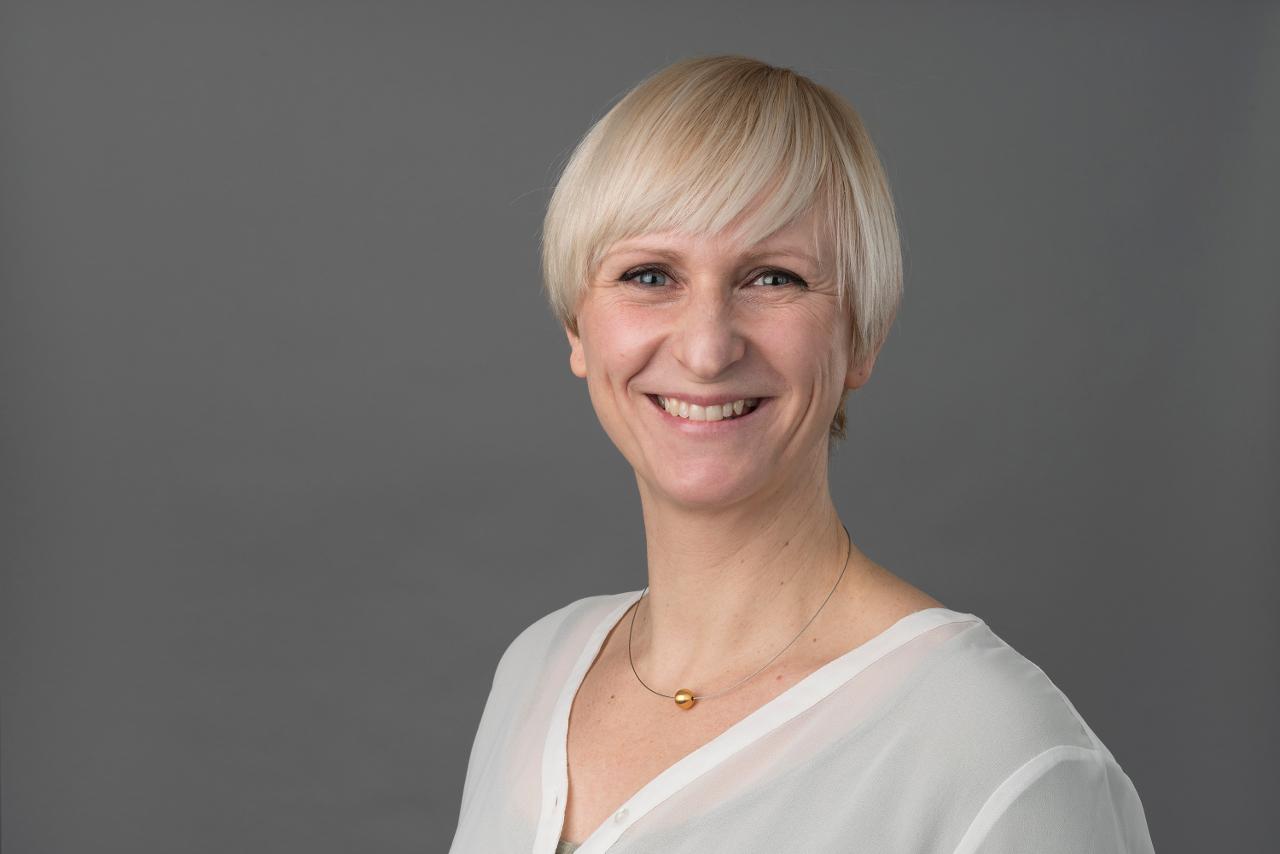 Antje Beckmann steht vor einem grauen Hintergrund und lächelt in die Kamera. Sie trägt kurze hellblonde Haare und eine weiße Bluse.